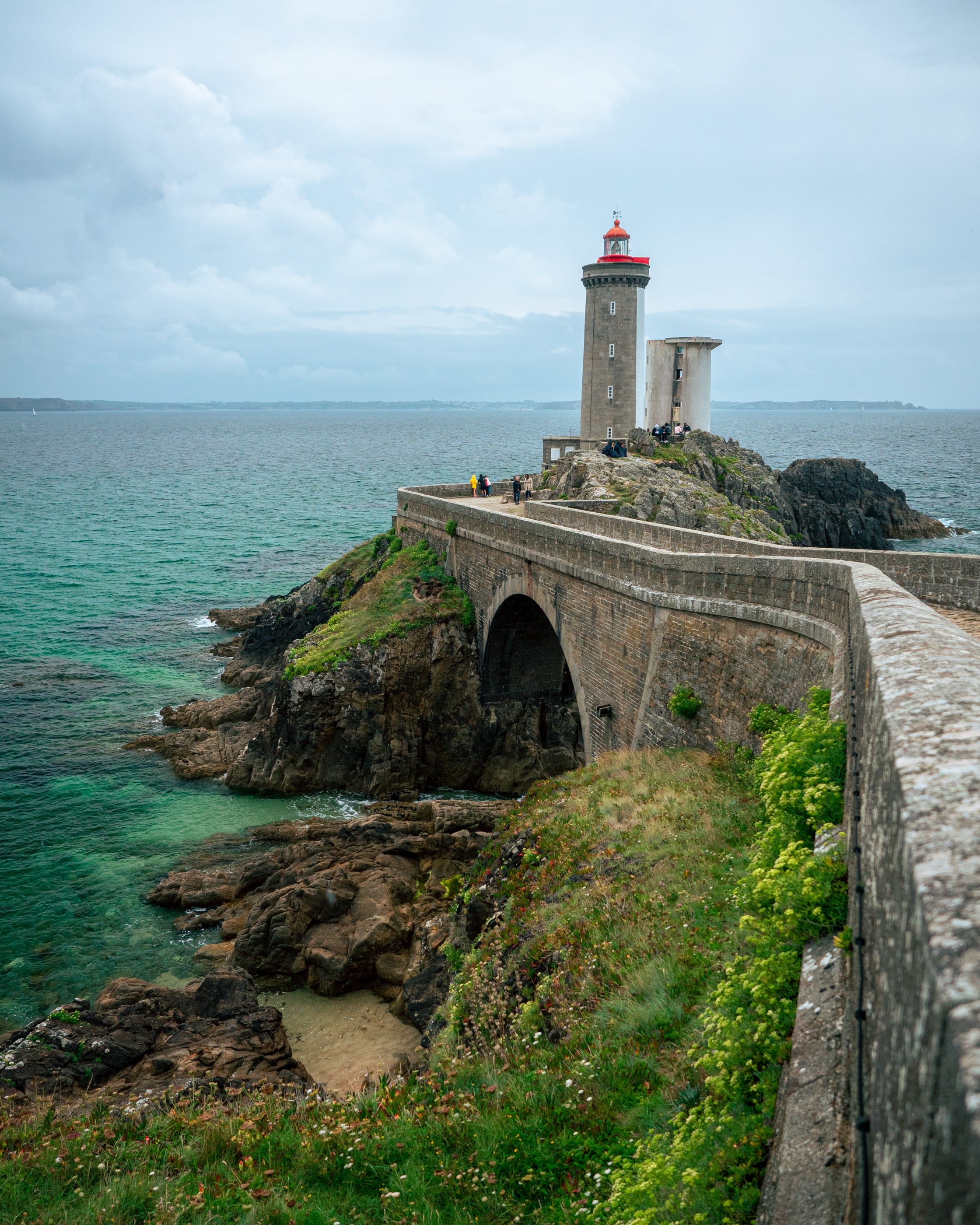 Un phare au sommet rouge relié à la côte par un pont de pierre, la mer turquoise autour des rochers.