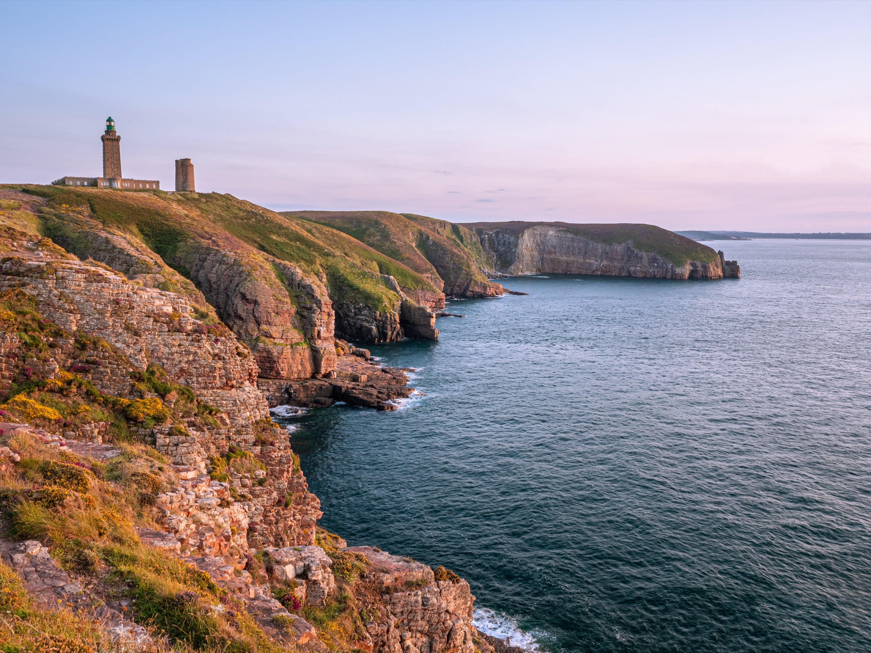 Le phare domine des falaises de grès rose couvertes de lande, la mer bleu sombre en contrebas sous un ciel rosé.