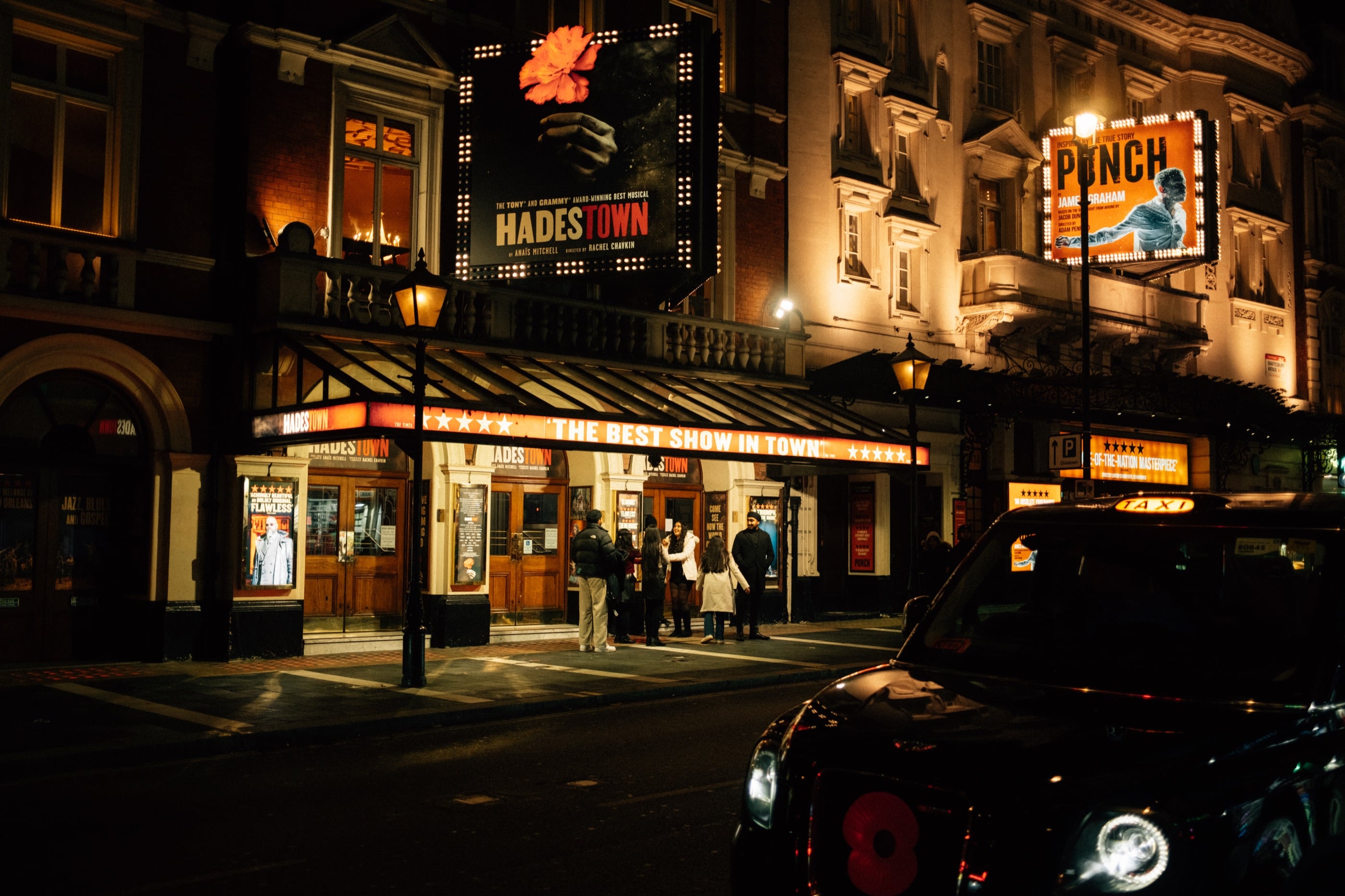 Façades de théâtres illuminées la nuit, grandes affiches lumineuses au-dessus des entrées, un taxi noir dans l'ombre au premier plan.