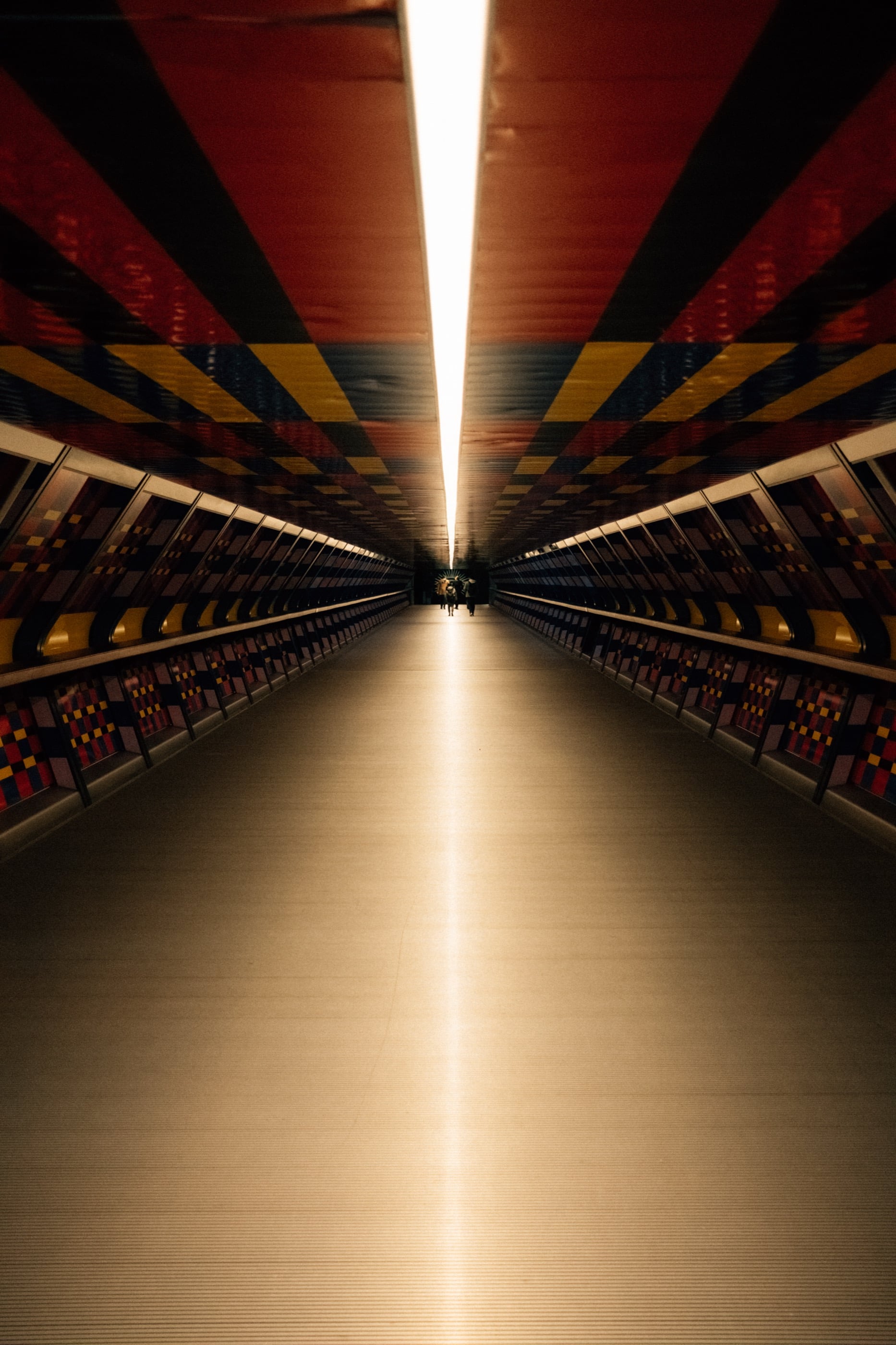 Un long couloir symétrique au plafond rayé de rouge, noir et jaune, une bande de lumière filant vers le point de fuite.