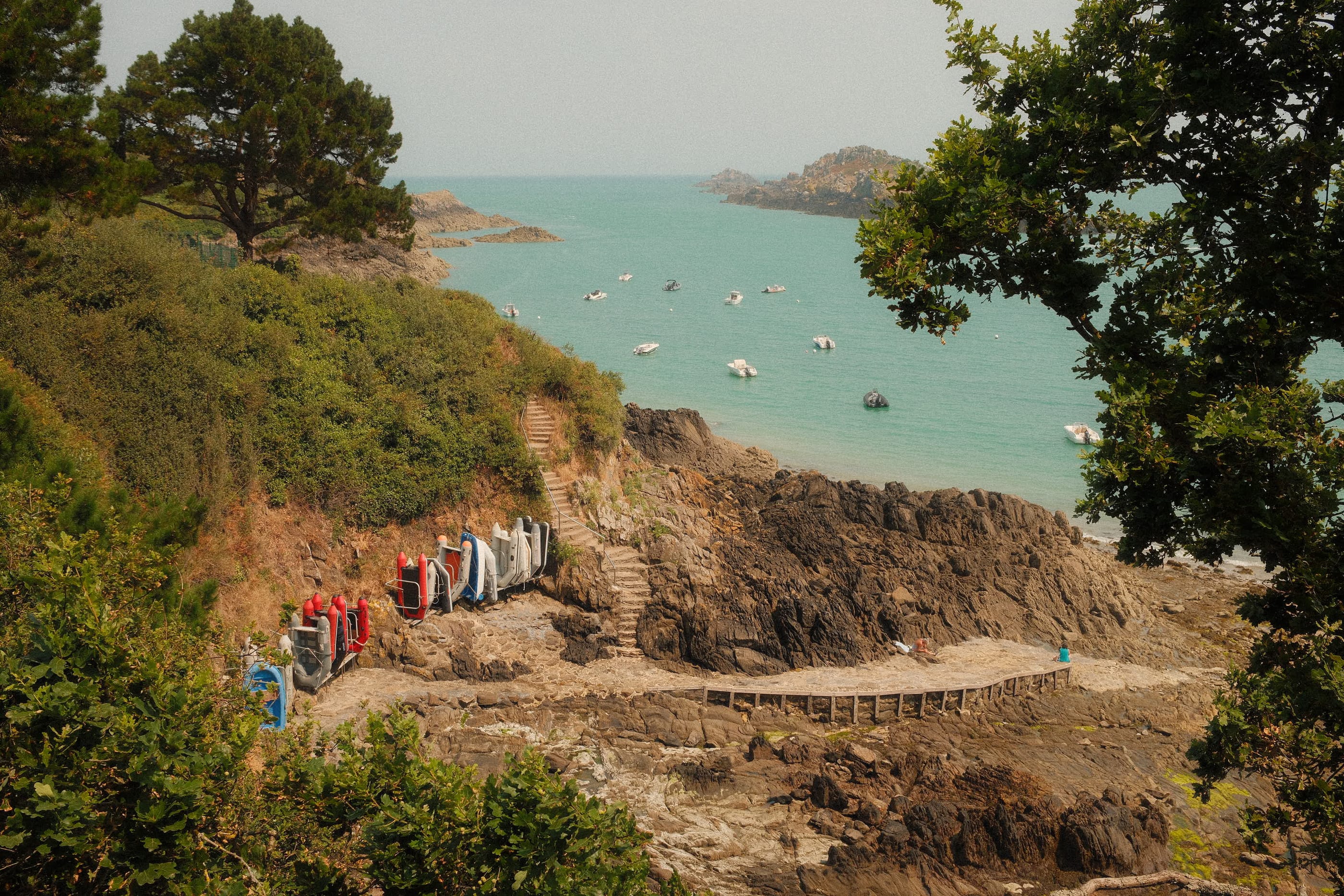 Une petite anse aux eaux turquoise vue d'en haut entre les arbres, des kayaks colorés rangés près d'un escalier, bateaux au mouillage.