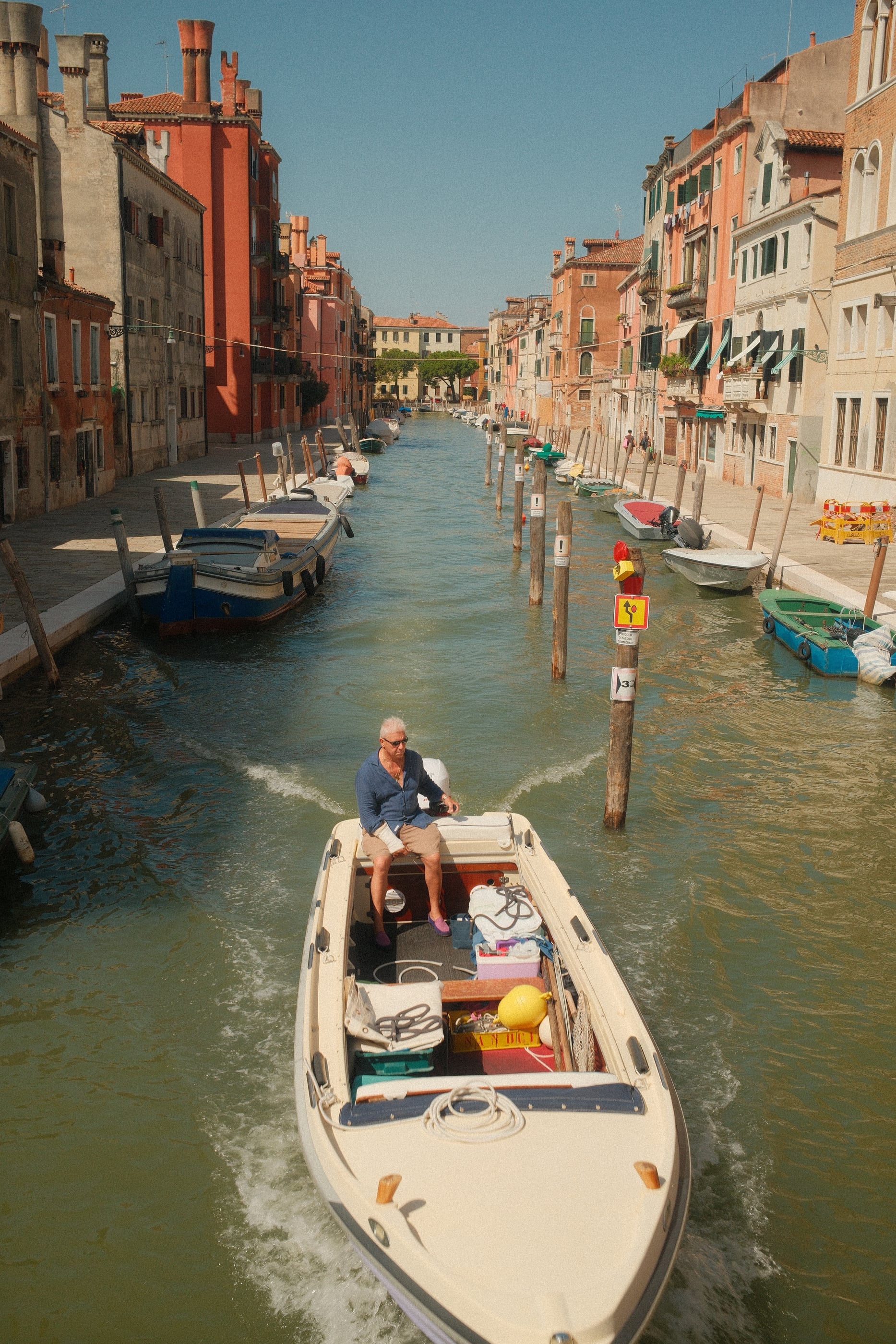 Un homme pilote un bateau à moteur crème sur un canal bordé de façades roses et ocre, sous un ciel bleu.