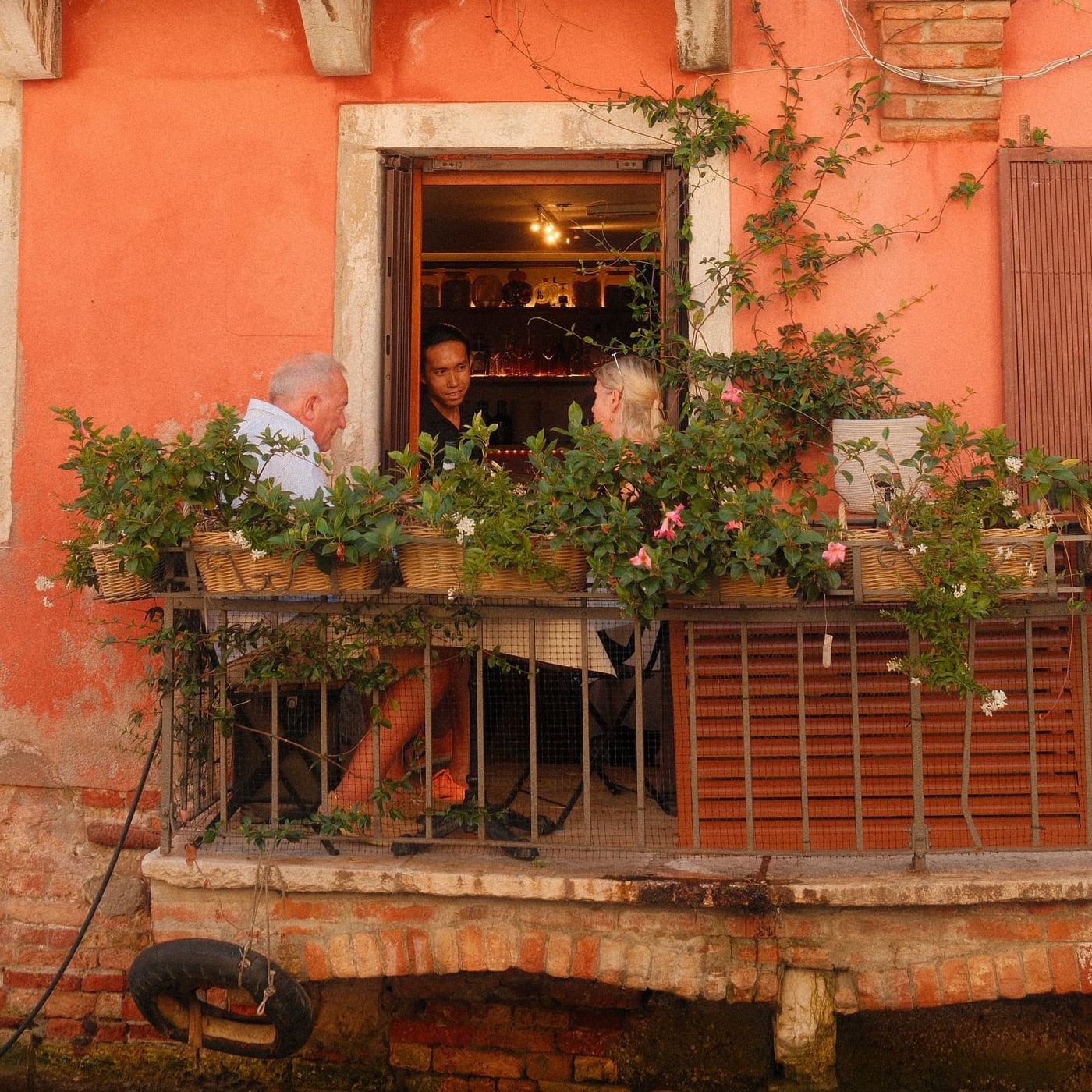 Un couple dîne sur un petit balcon fleuri accroché à une façade orangée, un serveur à la fenêtre, au-dessus de l'eau.
