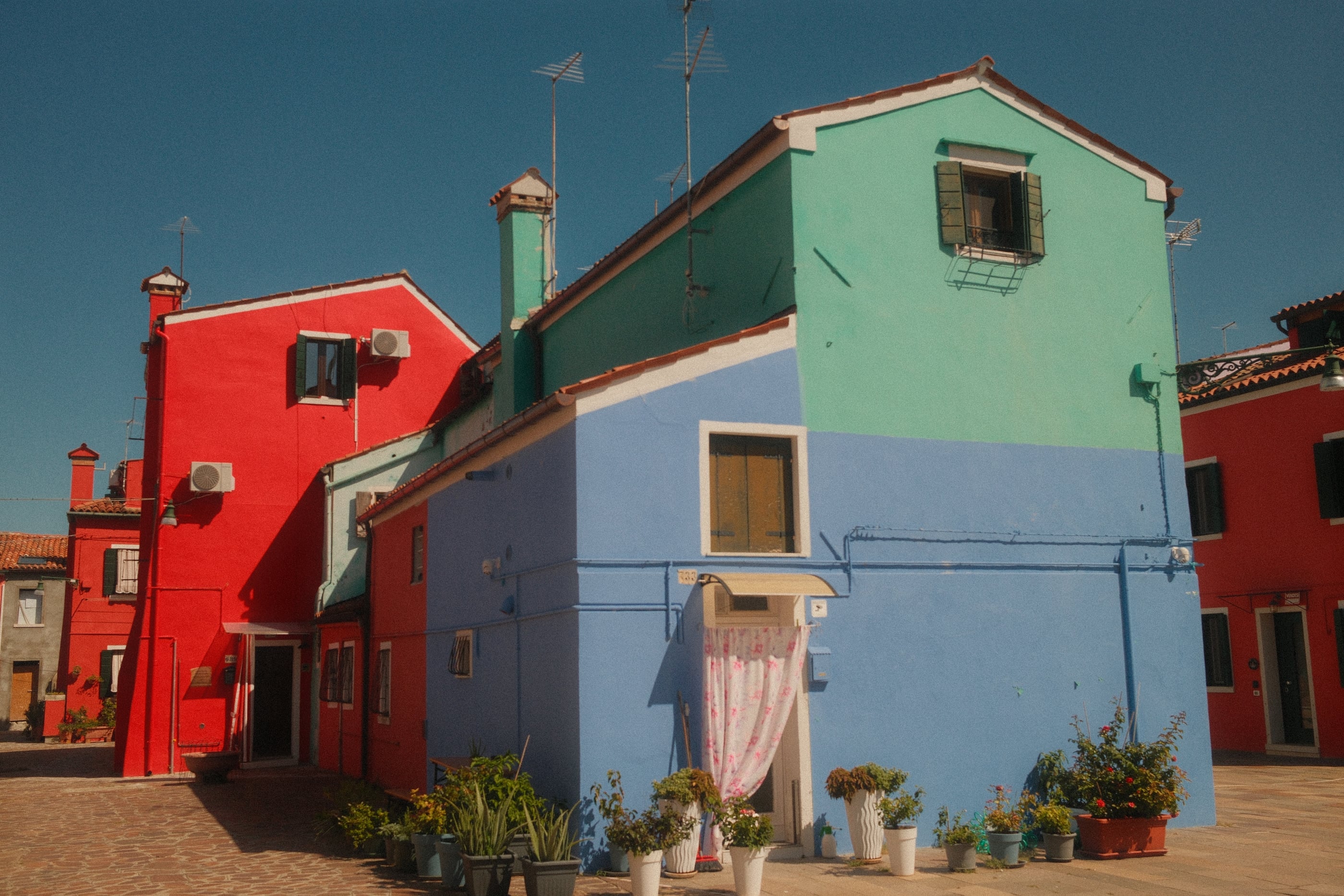 Maisons peintes en rouge, bleu et vert d'eau autour d'une placette garnie de pots de fleurs, sous un ciel bleu profond.
