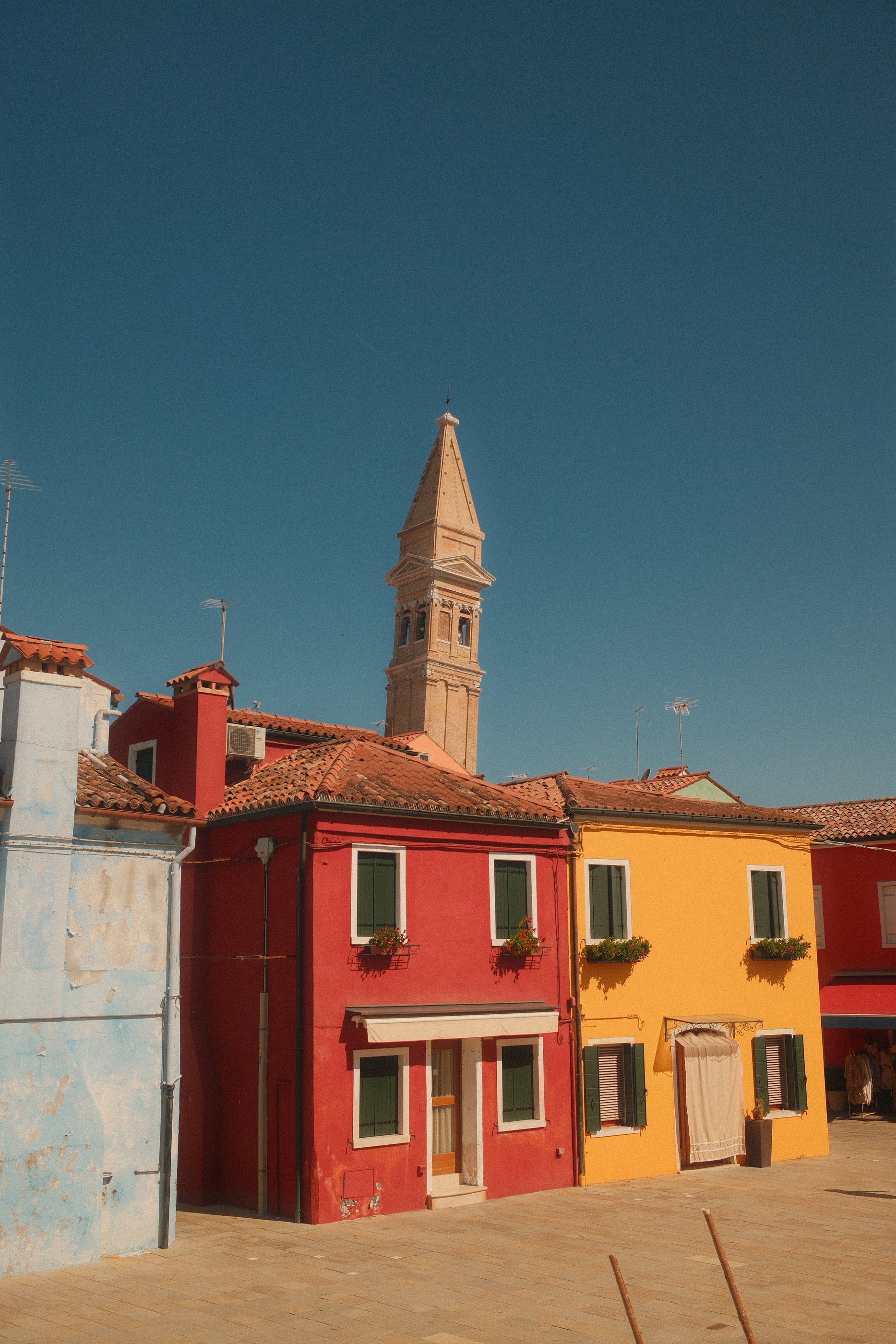 Un clocher incliné dépasse des toits de maisons rouge et jaune, sur un ciel bleu sans nuage.