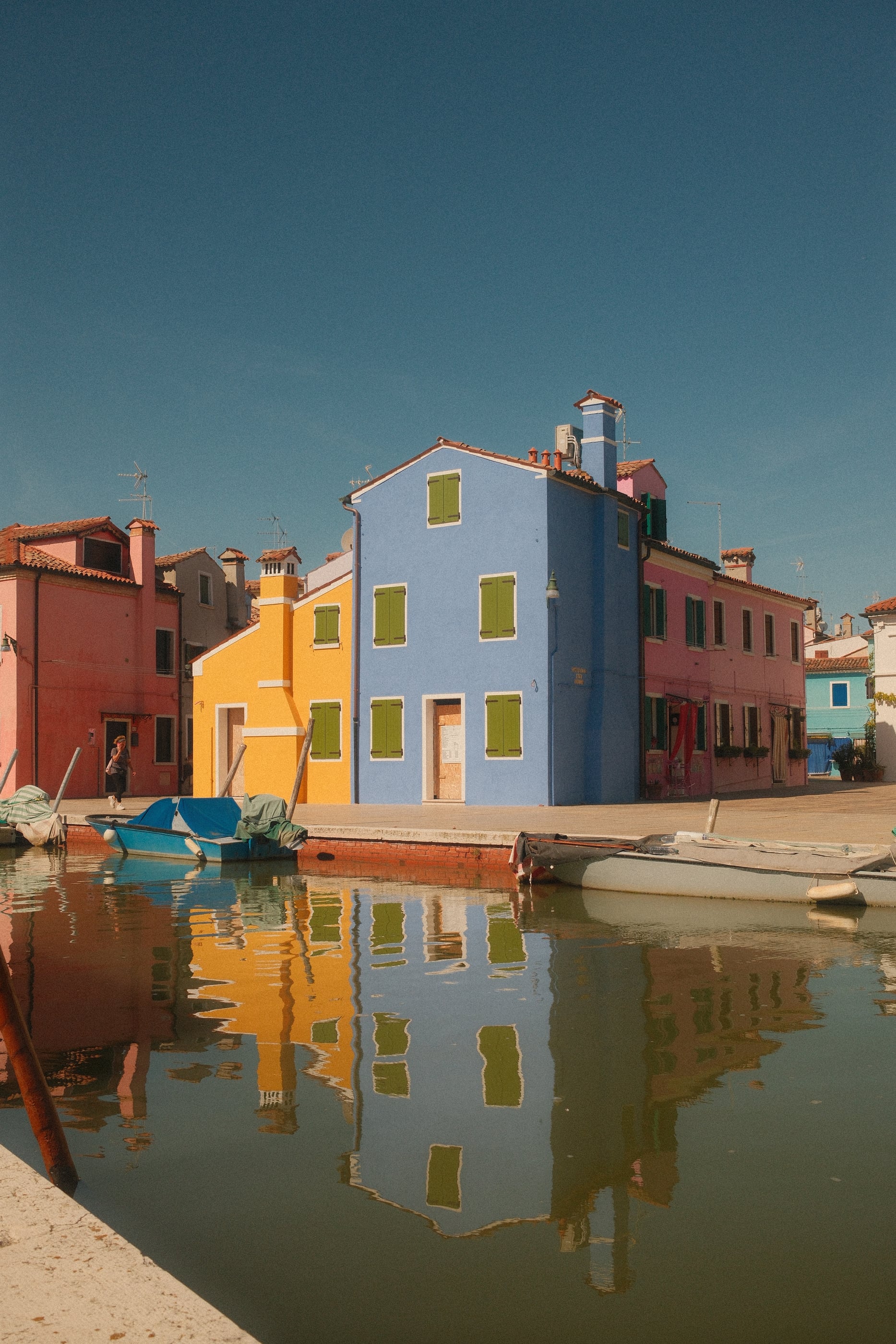 Maisons bleue, jaune et rose aux volets verts reflétées dans l'eau calme d'un canal, des barques amarrées au quai.