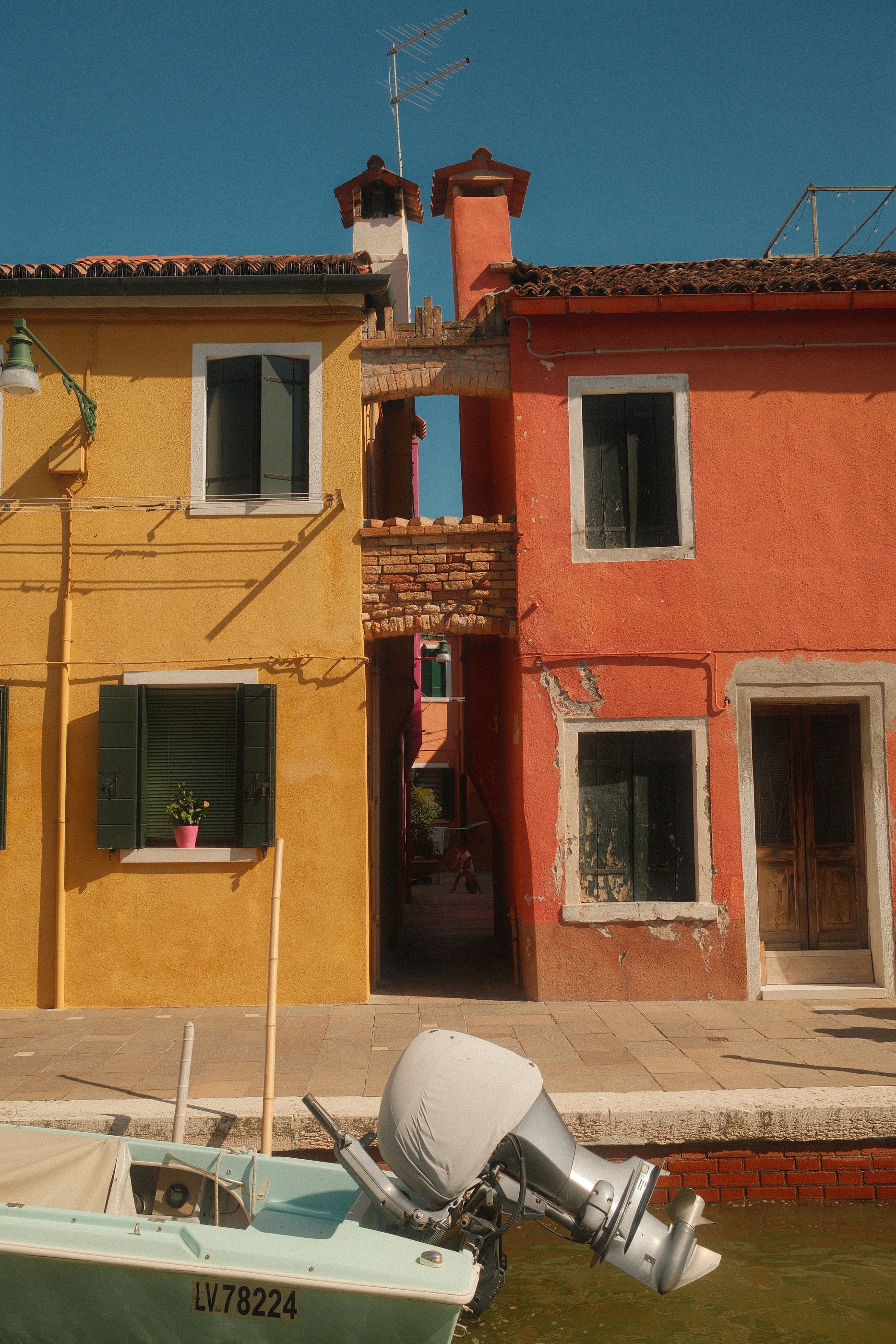 Une maison jaune et une maison corail séparées par un étroit passage, le moteur d'une barque au premier plan.