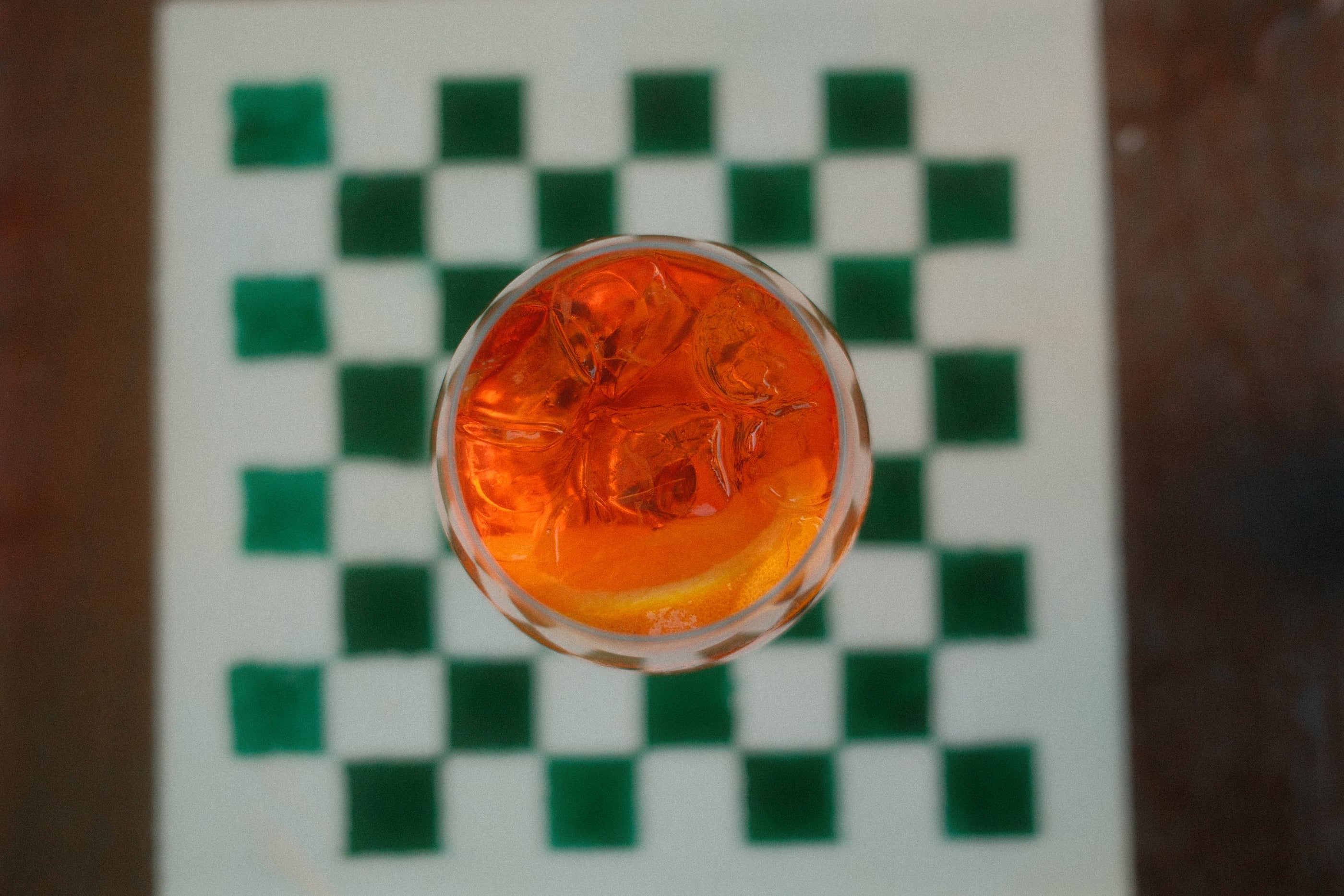 Vu du dessus, un verre orange avec glaçons et tranche d'orange posé sur une nappe à damier vert et blanc.