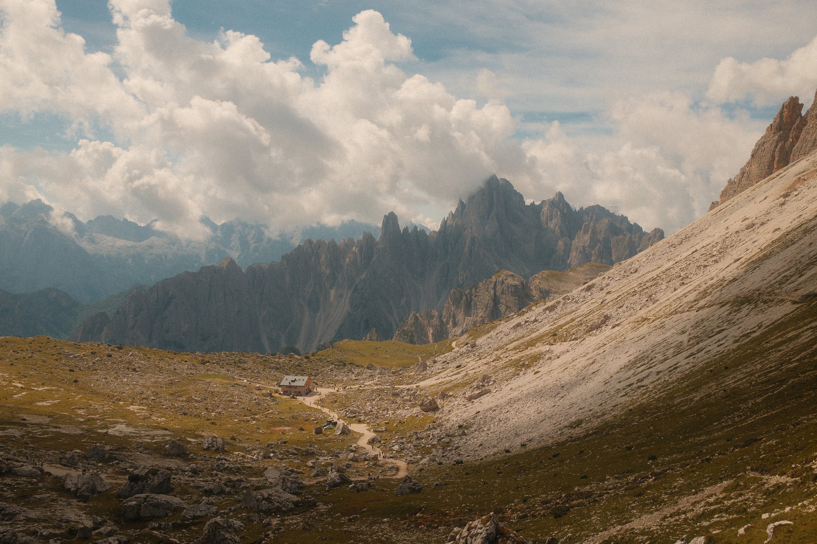Un sentier mène à un refuge isolé au creux d'une vallée d'éboulis, des crêtes dentelées sous de gros nuages.