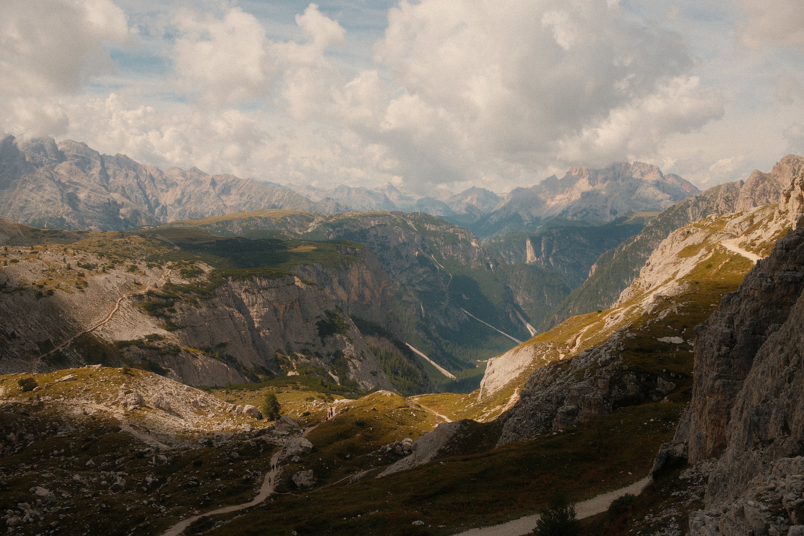 Une vallée profonde vue depuis les hauteurs, des sentiers dans l'herbe rase et des chaînes de montagnes sous les nuages.