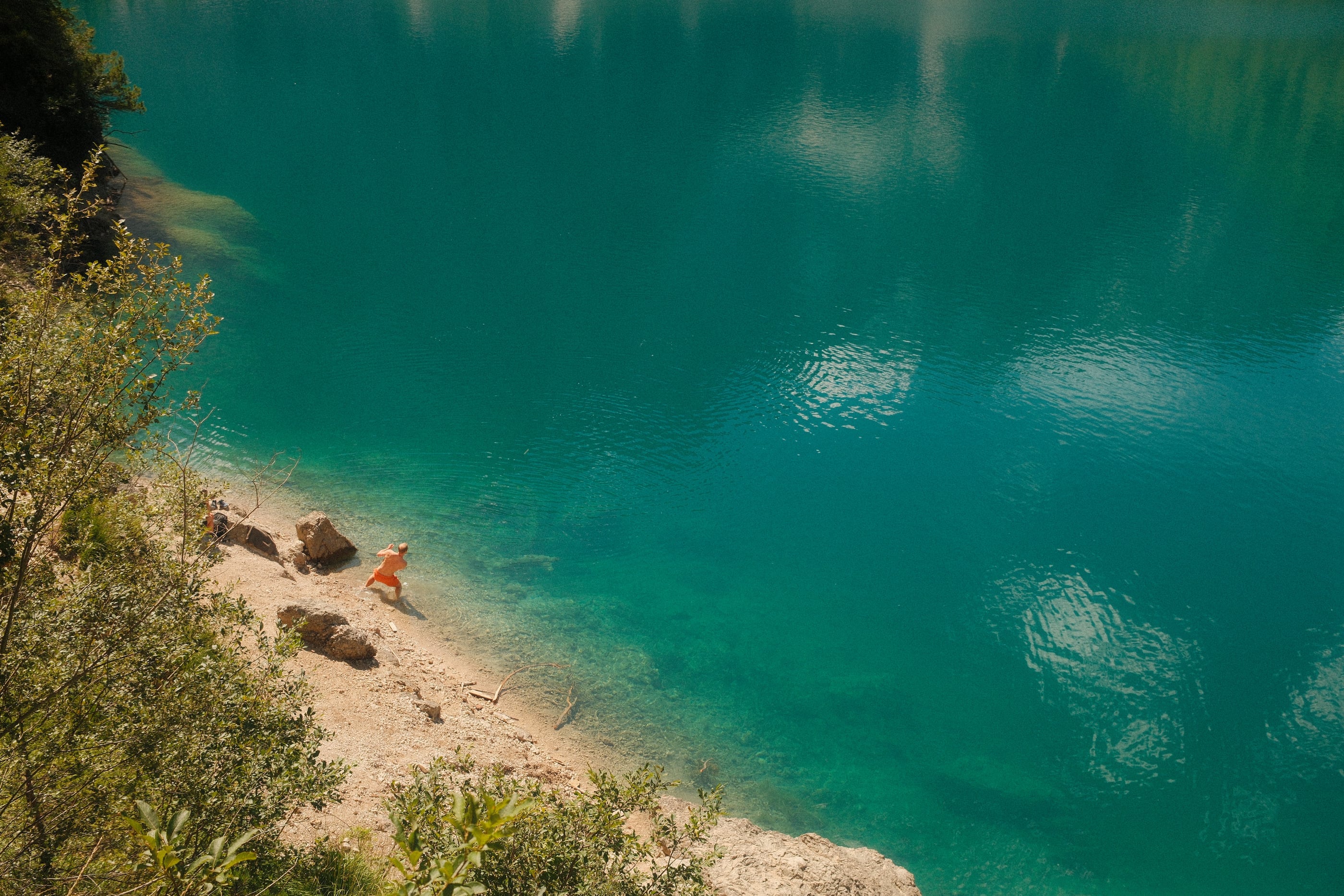 Vue plongeante sur une eau bleu-vert transparente, une baigneuse en orange sur la rive de galets clairs.