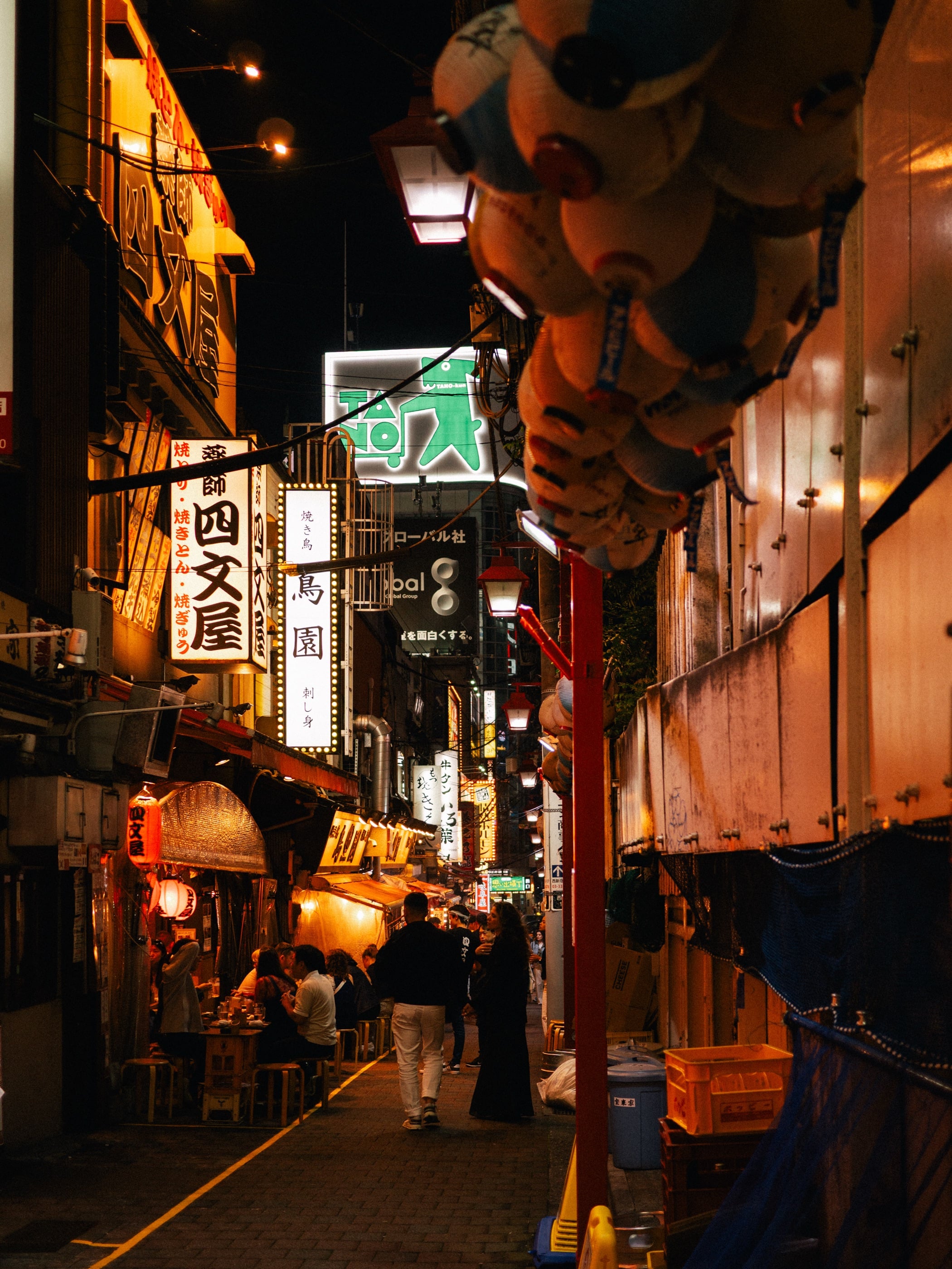 Une ruelle étroite la nuit, enseignes lumineuses en japonais et lampions, des clients attablés devant de petits bars.