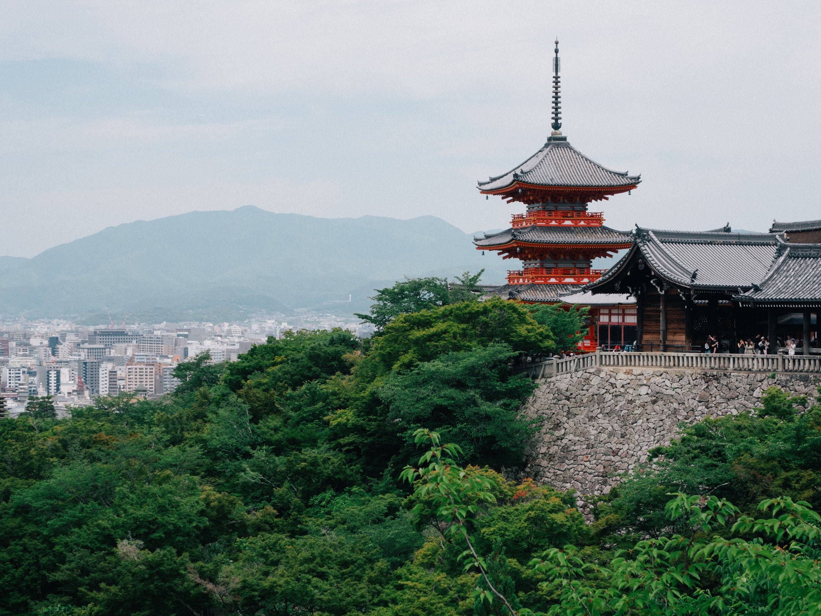 Une pagode rouge à trois étages et les toits du temple au-dessus d'une colline boisée, la ville et les montagnes au loin.