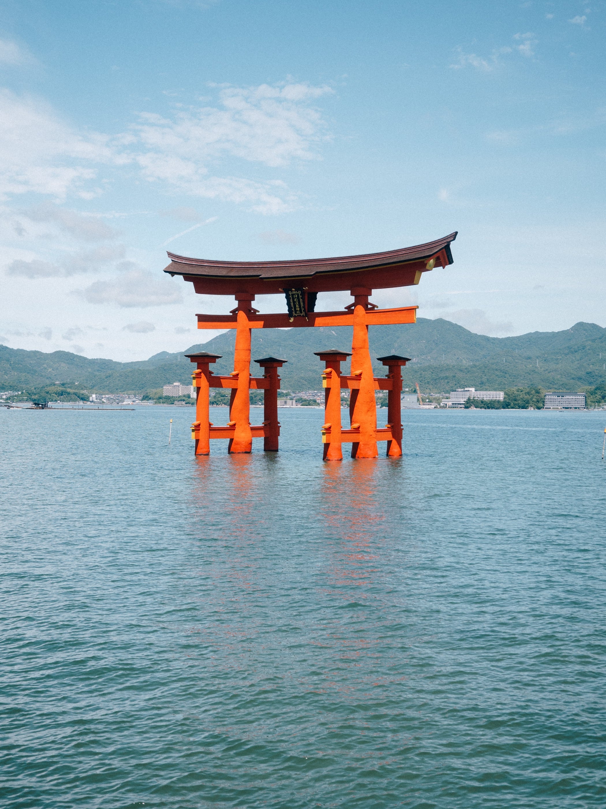 Le grand portique orange se dresse dans la mer, les montagnes boisées et un ciel bleu légèrement nuageux derrière.
