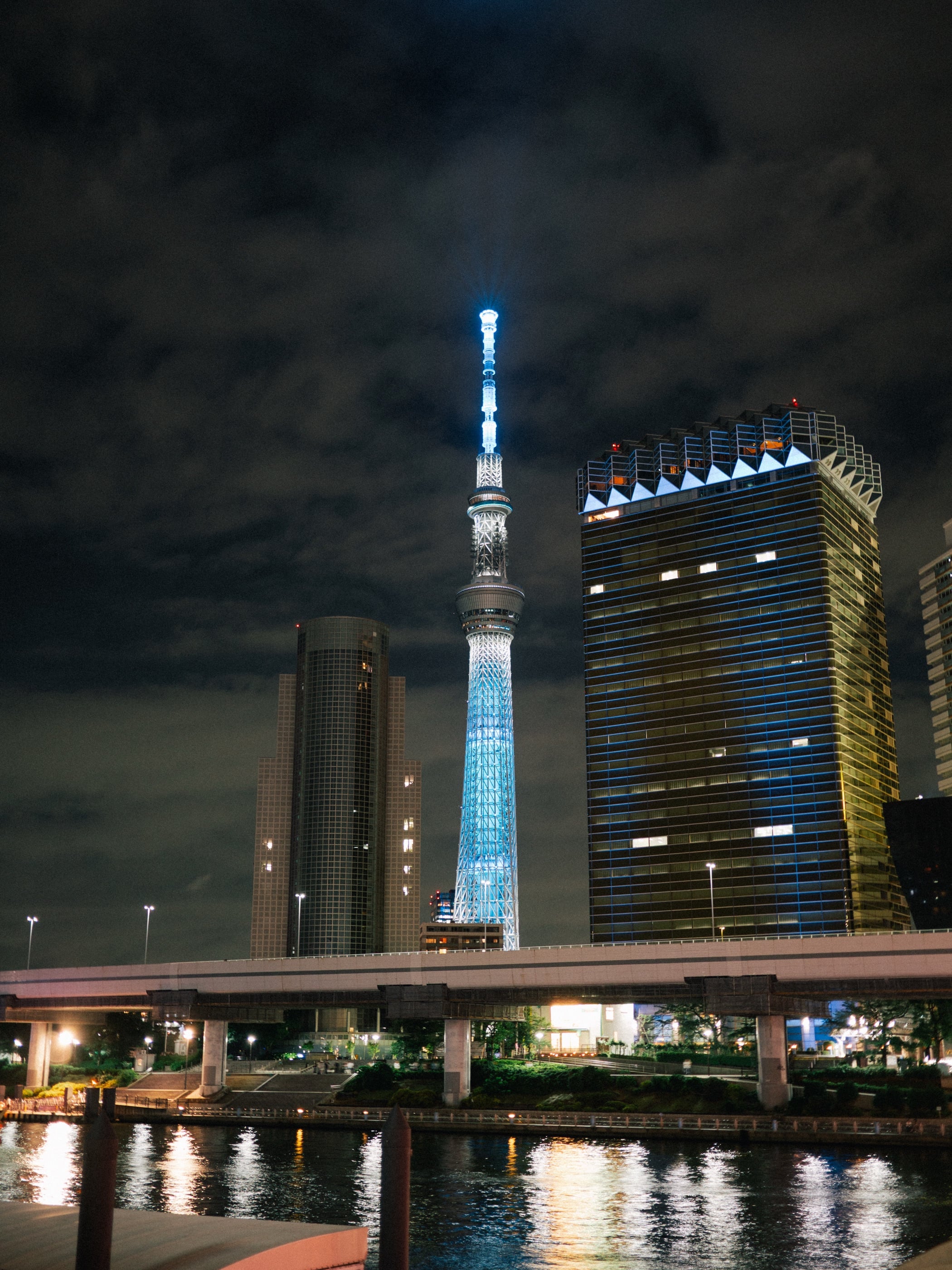 La tour illuminée de bleu s'élance entre deux immeubles sous un ciel nocturne nuageux, ses lumières reflétées dans la rivière.
