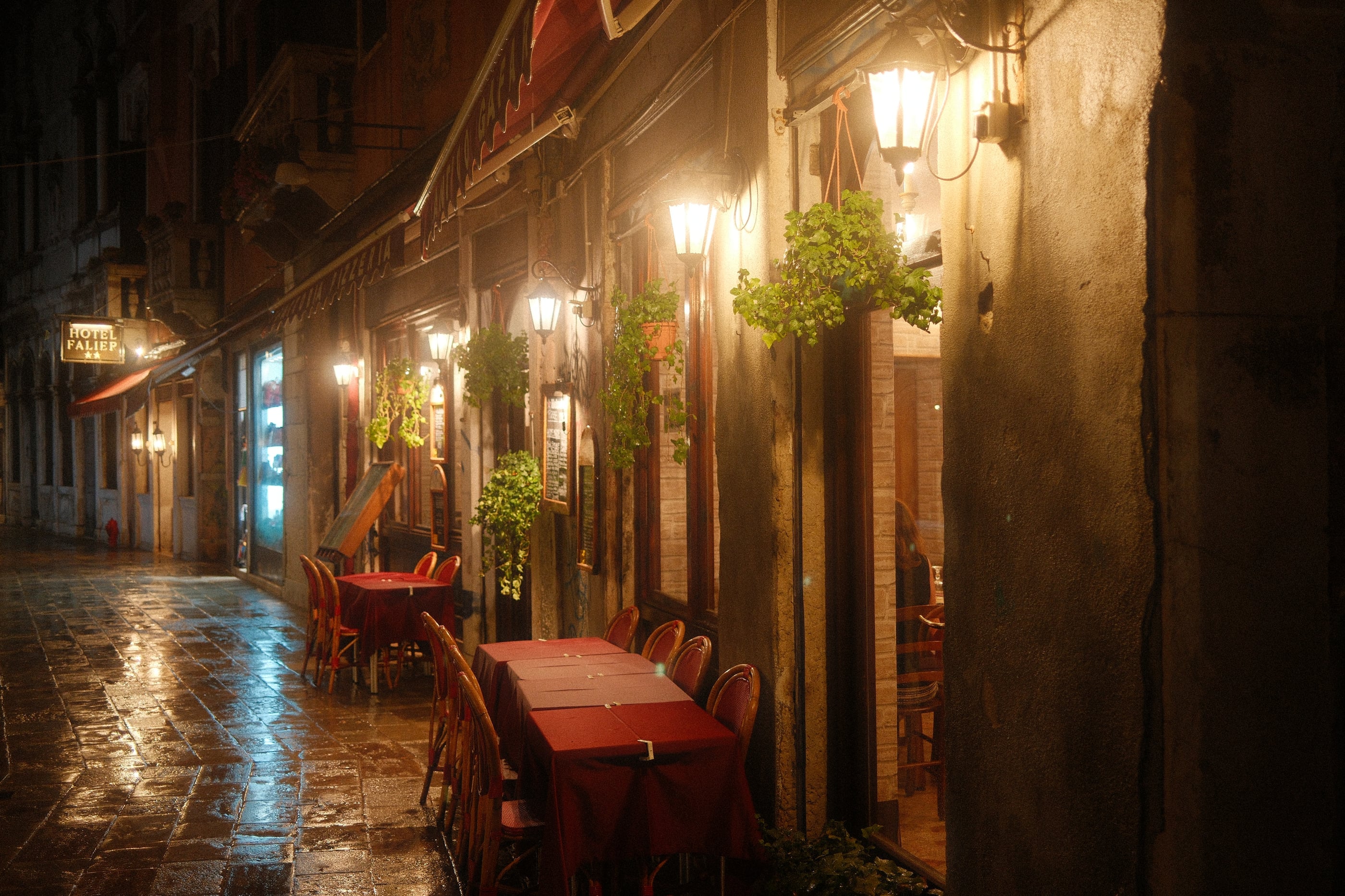 Une terrasse de restaurant aux nappes rouges éclairée par des lanternes, les pavés mouillés luisant dans une ruelle la nuit.