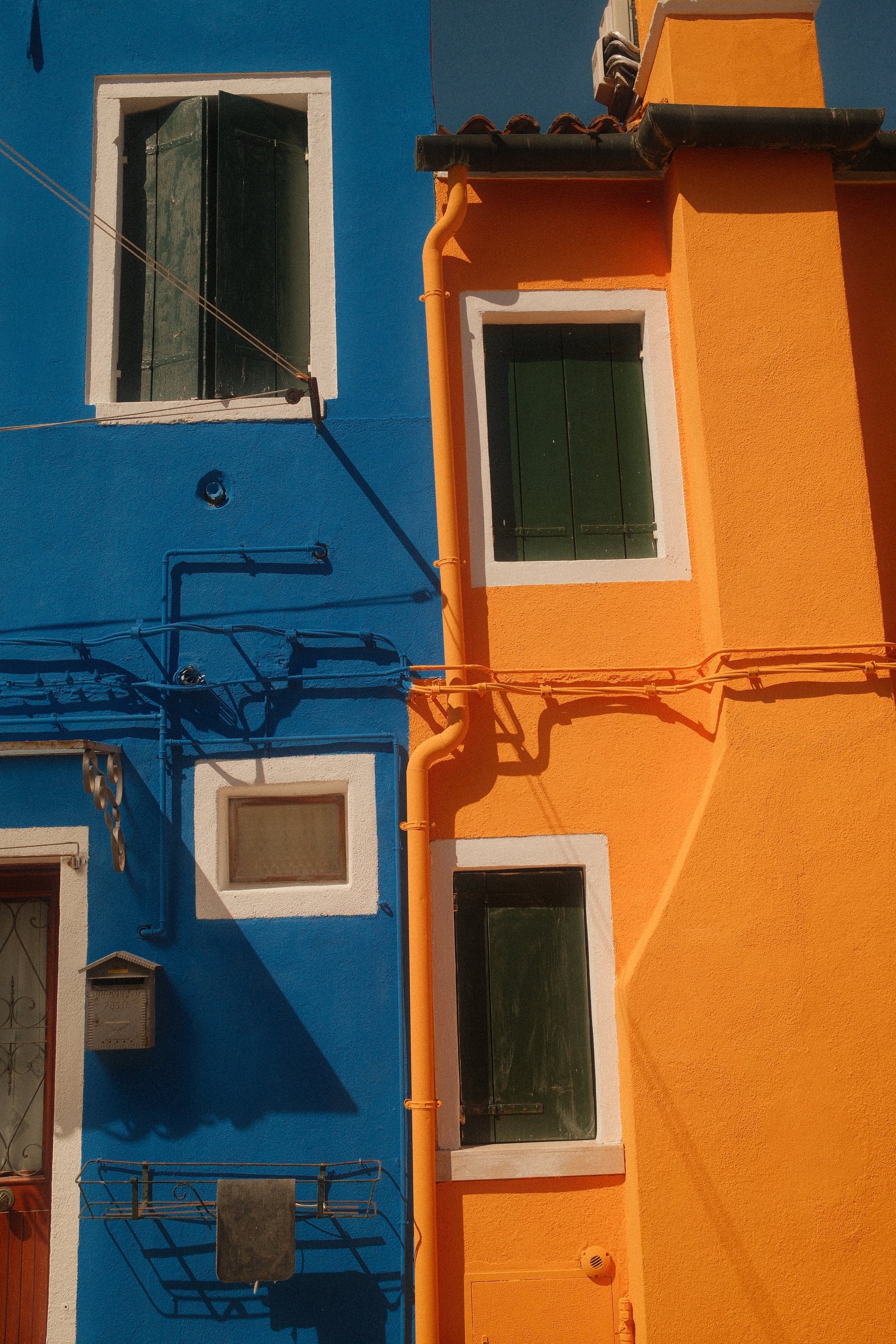Deux façades accolées, l'une bleu vif, l'autre orange, avec leurs fenêtres aux volets verts et leurs gouttières.