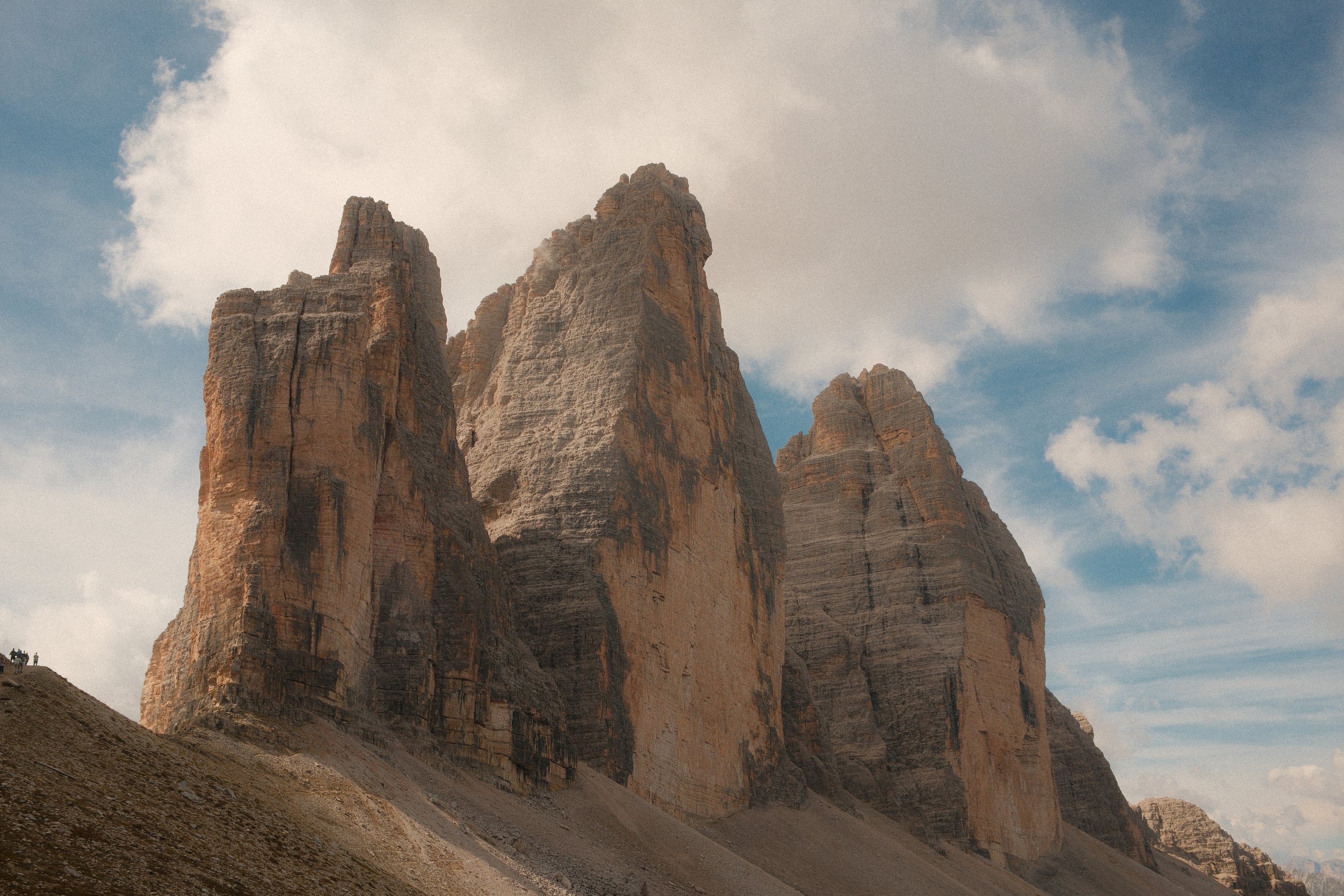 Trois tours de roche ocre se dressent au-dessus d'un éboulis, sous un ciel bleu et de gros nuages blancs.