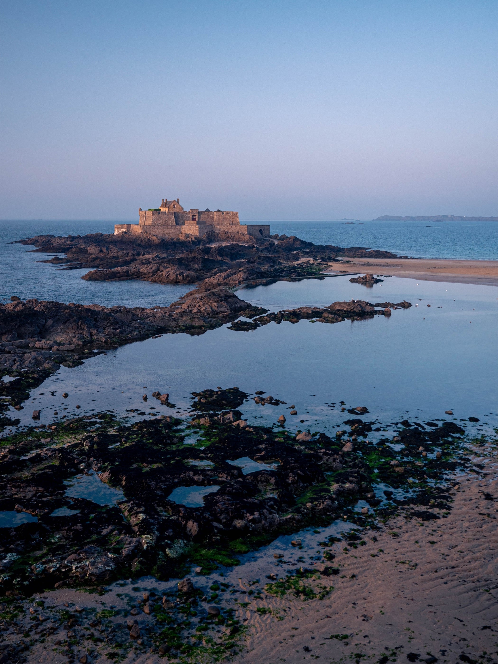 Le fort se dresse sur son îlot rocheux à marée basse, flaques et rochers couverts d'algues au premier plan.