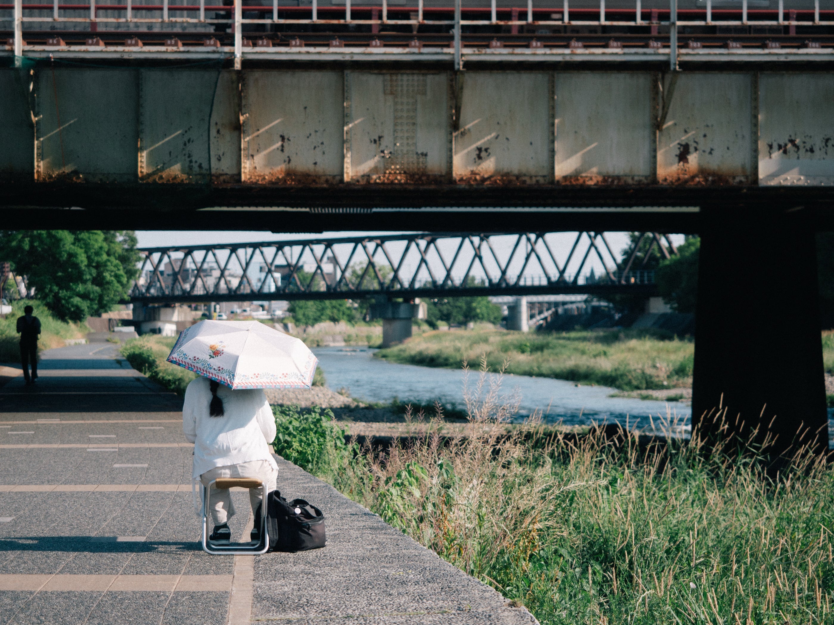Sous un pont ferroviaire rouillé, une personne assise sur un tabouret s'abrite sous une ombrelle blanche au bord de la rivière.